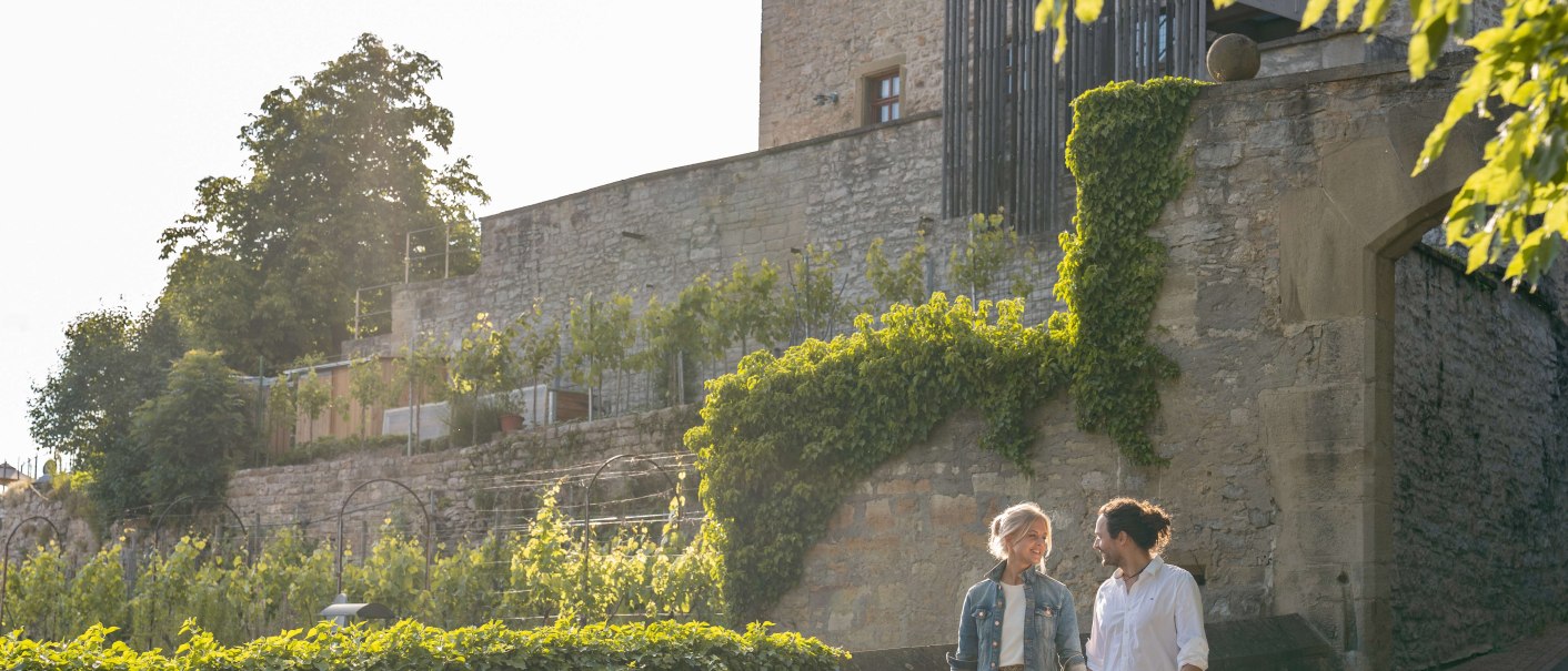 Ein Paar spaziert Hand in Hand vor einem alten Steinhaus mit Efeu und Weinreben im Sonnenlicht., © SMG, Martina Denker