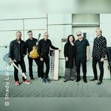 Sechs Personen stehen mit Musikinstrumenten vor einem Gebäude. Sie tragen überwiegend schwarze Kleidung und lächeln in die Kamera., © links im Bild