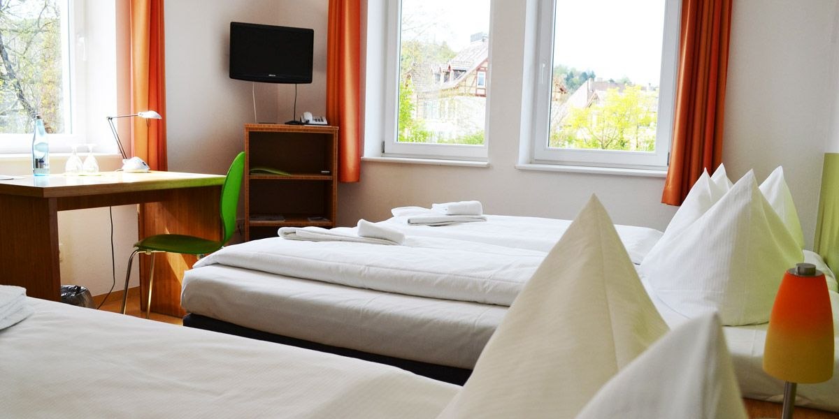 Helles Zimmer mit zwei Betten, Schreibtisch, gr&uuml;nem Stuhl und Fernseher. Gro&szlig;e Fenster mit orangefarbenen Vorh&auml;ngen., &copy; ECOINN Hotel am Campus