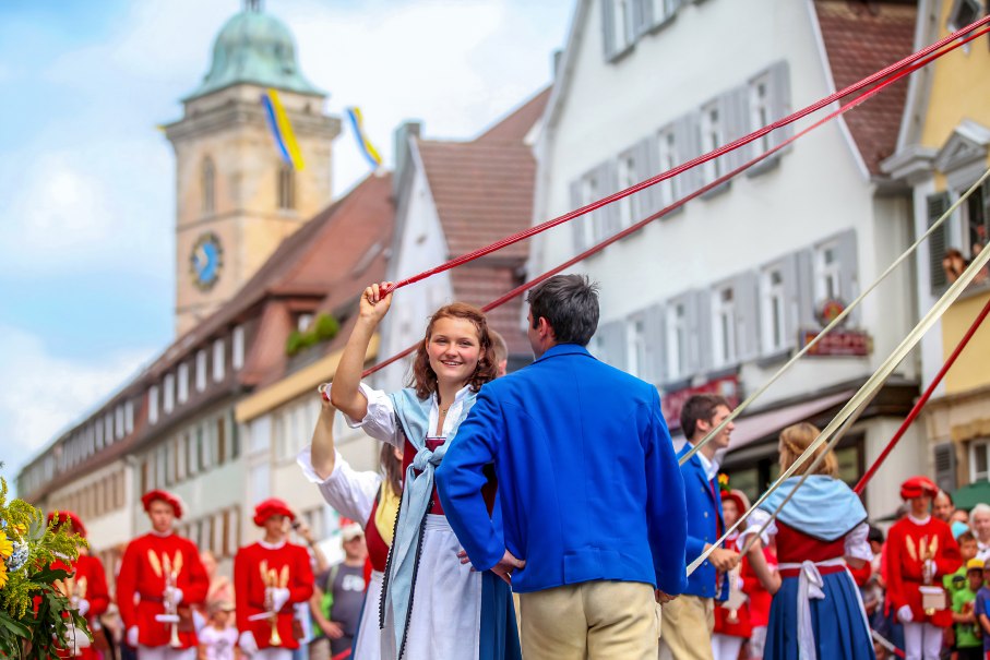 Menschen in traditioneller Kleidung tanzen mit Bändern auf einem Fest in einer historischen Stadt. Im Hintergrund sind Zuschauer und ein Turm zu sehen., © D. Jüptner
