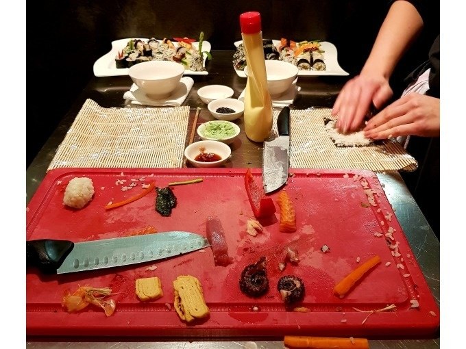 Sushi-Zubereitung mit Zutaten auf Schneidebrett, Bambusmatte und fertigen Sushirollen im Hintergrund., &copy; Kultweine-Kochwelten-Kunst&uuml;cke - Kochschule Sonja Lenz