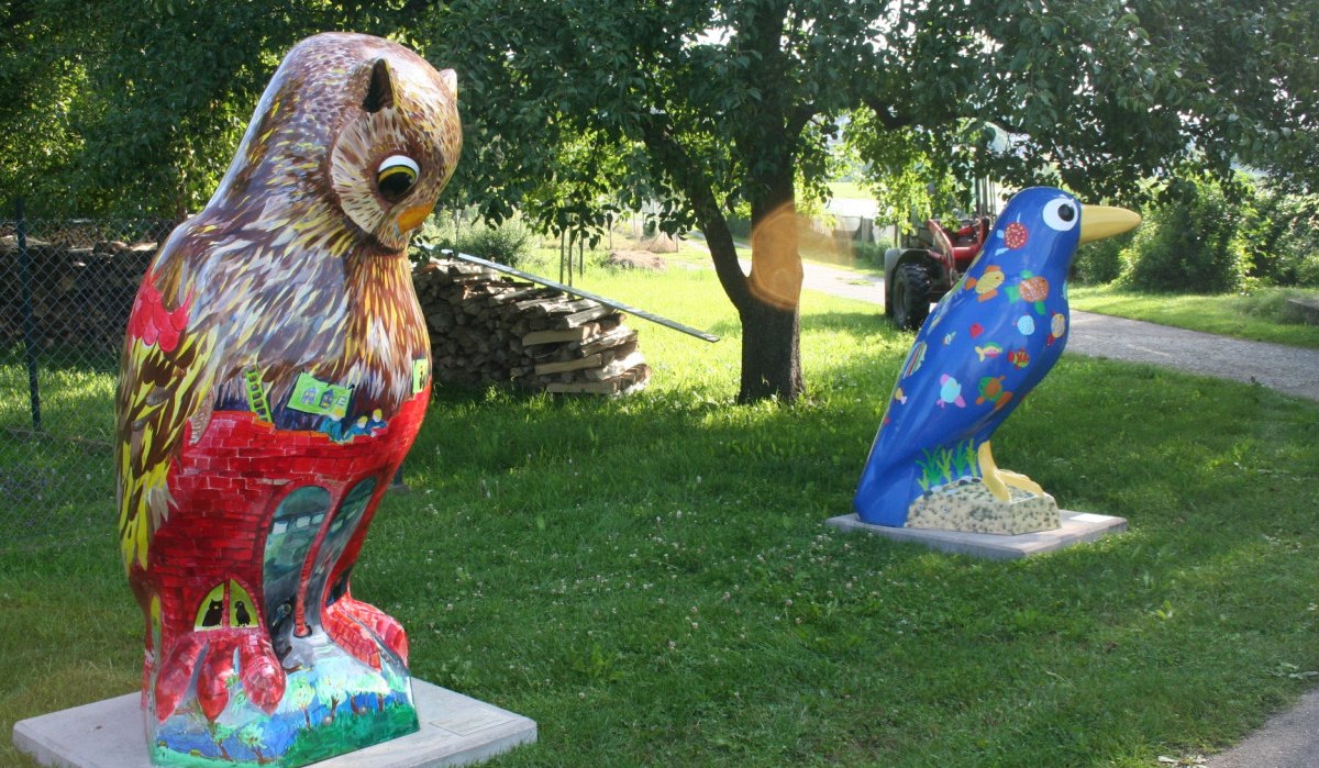 Zwei bunte Vogelskulpturen, eine Eule und ein blauer Vogel, stehen auf einer Wiese vor Bäumen., © Natur.Nah. Schönbuch & Heckengäu