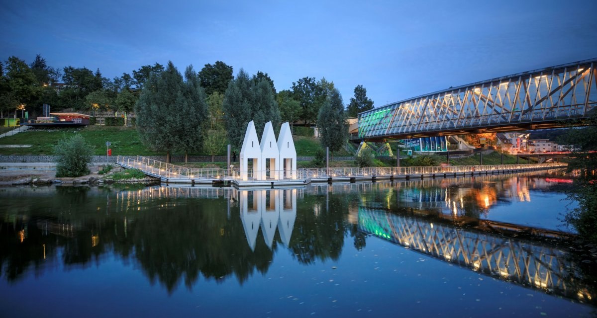 Moderne Brücke und dreieckige Gebäude am Flussufer bei Abenddämmerung, mit Spiegelung im Wasser., © Stoppel Moderne Brücke und dreieckige Gebäude am Flussufer bei Abenddämmerung, mit Spiegelung im Wasser., © Stoppel