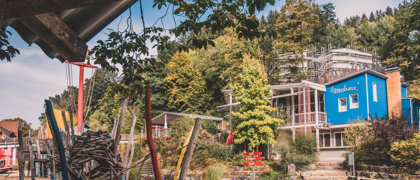 Spielplatz mit buntem Klettergerüst, Holzspänen und einem blauen Gebäude im Hintergrund, umgeben von Bäumen., © Stadt Welzheim Spielplatz mit buntem Klettergerüst, Holzspänen und einem blauen Gebäude im Hintergrund, umgeben von Bäumen., © Stadt Welzheim