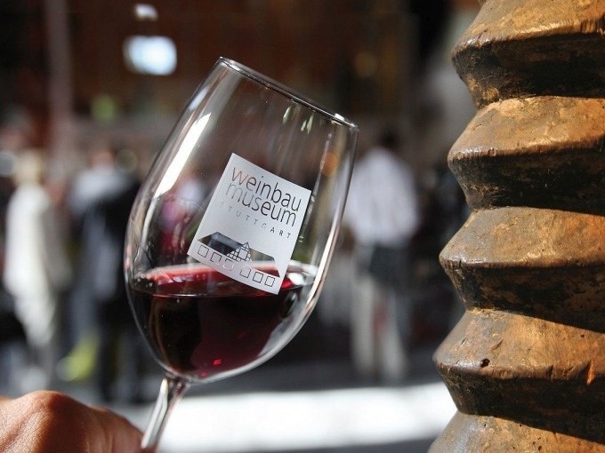Ein Weinglas mit dem Logo des Weinbaumuseums Stuttgart, gef&uuml;llt mit Rotwein, vor einem unscharfen Hintergrund mit Menschen., &copy; Stuttgart Marketing GmbH