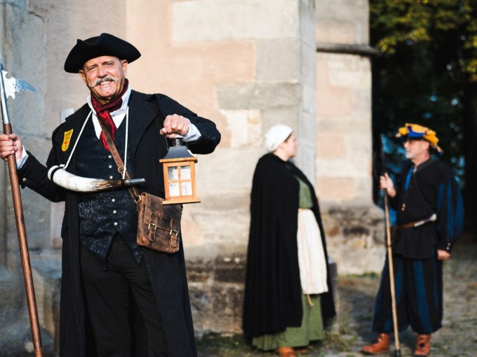 Person in historischer Kleidung mit Laterne und Hellebarde, zwei weitere Personen im Hintergrund vor einem alten Gebäude., © Stadt Nürtingen