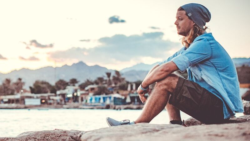 Ein Mann mit M&uuml;tze und Hemd sitzt nachdenklich auf einem Felsen am Meer. Im Hintergrund sind Berge und ein Dorf zu sehen, w&auml;hrend die Sonne untergeht., &copy; Stadthalle Leonberg