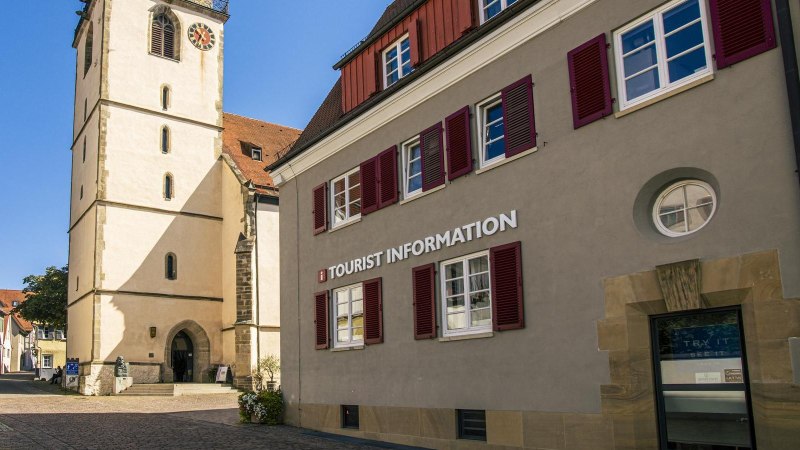 Gebäude mit der Aufschrift 'Tourist Information' neben einer Kirche mit Turm und roten Fensterläden, bei sonnigem Wetter., © Stuttgart-Marketing GmbH, Sarah Schmid