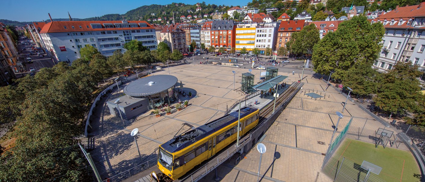 Luftaufnahme des Marienplatzes in Stuttgart. Eine gelbe Straßenbahn fährt vorbei, umgeben von Bäumen und Gebäuden. Der Platz ist belebt und sonnig., © Stuttgart-Marketing GmbH, Achim Mende Luftaufnahme des Marienplatzes in Stuttgart. Eine gelbe Straßenbahn fährt vorbei, umgeben von Bäumen und Gebäuden. Der Platz ist belebt und sonnig., © Stuttgart-Marketing GmbH, Achim Mende