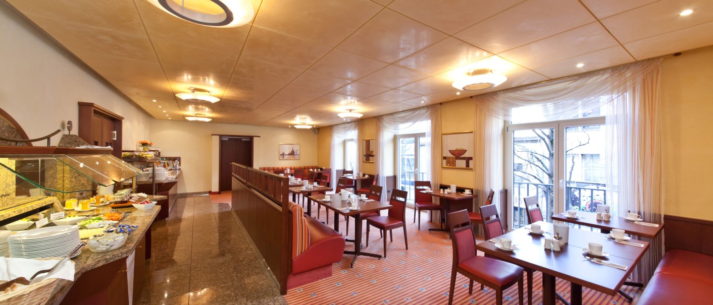 Eleganter Frühstücksraum mit roten Stühlen, gedeckten Tischen und einem Buffet. Große Fenster lassen viel Licht herein., © Novum Boulevard Stuttgart City