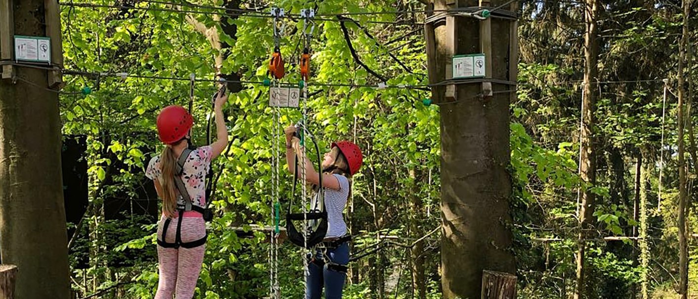 Zwei Personen mit roten Helmen klettern im Skypark Kletterwald in Schwäbisch Gmünd. Sie sind mit Sicherheitsgurten gesichert und von Bäumen umgeben., © SKYPARK – epia-store.de GbR