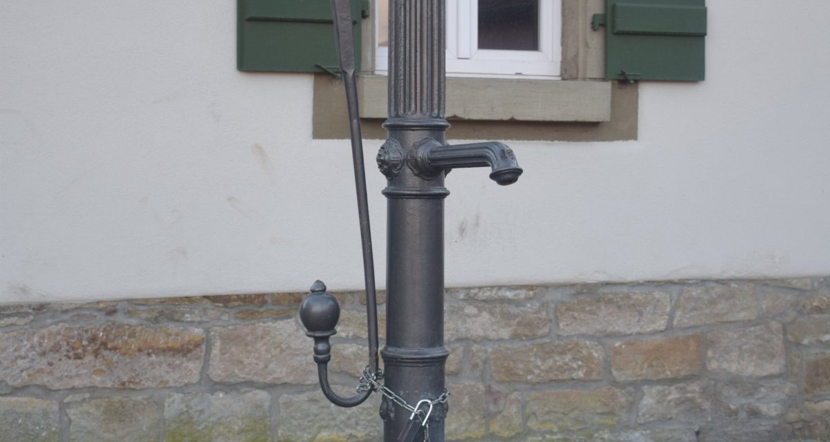 Ein alter Pumpbrunnen aus Metall steht vor einem Gebäude mit grünen Fensterläden und einem steinernen Sockel., © Land der 1000 Hügel - Kraichgau-Stromberg Ein alter Pumpbrunnen aus Metall steht vor einem Gebäude mit grünen Fensterläden und einem steinernen Sockel., © Land der 1000 Hügel - Kraichgau-Stromberg