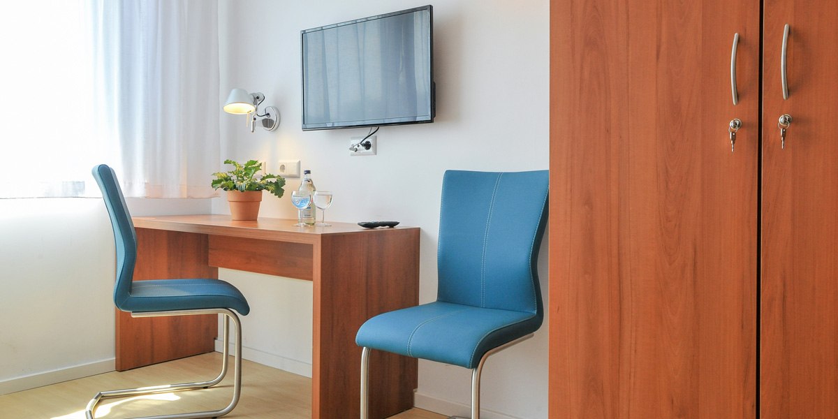 Ein schlichtes Hotelzimmer mit Schreibtisch, zwei blauen Stühlen, einem Fernseher an der Wand und einem Kleiderschrank., © Gästehaus ABZ Stuttgart Ein schlichtes Hotelzimmer mit Schreibtisch, zwei blauen Stühlen, einem Fernseher an der Wand und einem Kleiderschrank., © Gästehaus ABZ Stuttgart