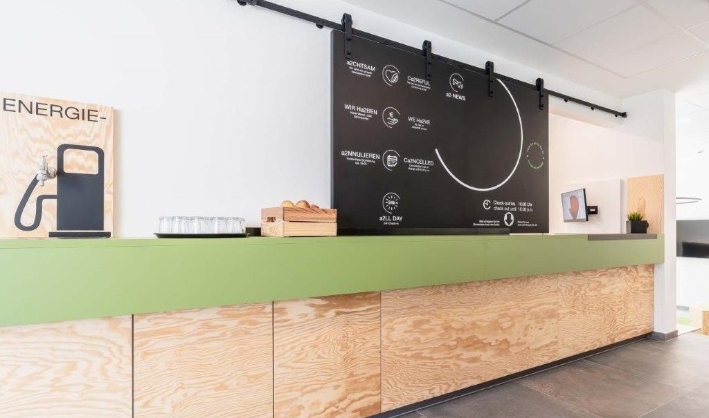 Moderne Rezeption mit grünem Tresen, Holzverkleidung, schwarzer Infotafel, Wasserstation und kleinen Pflanzen., © a2 HOTELS am Quadrium