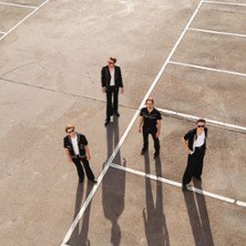 Vier Personen stehen auf einem leeren Parkplatz, ihre langen Schatten fallen auf den Boden. Sie tragen dunkle Kleidung und Sonnenbrillen., © links im Bild Vier Personen stehen auf einem leeren Parkplatz, ihre langen Schatten fallen auf den Boden. Sie tragen dunkle Kleidung und Sonnenbrillen., © links im Bild