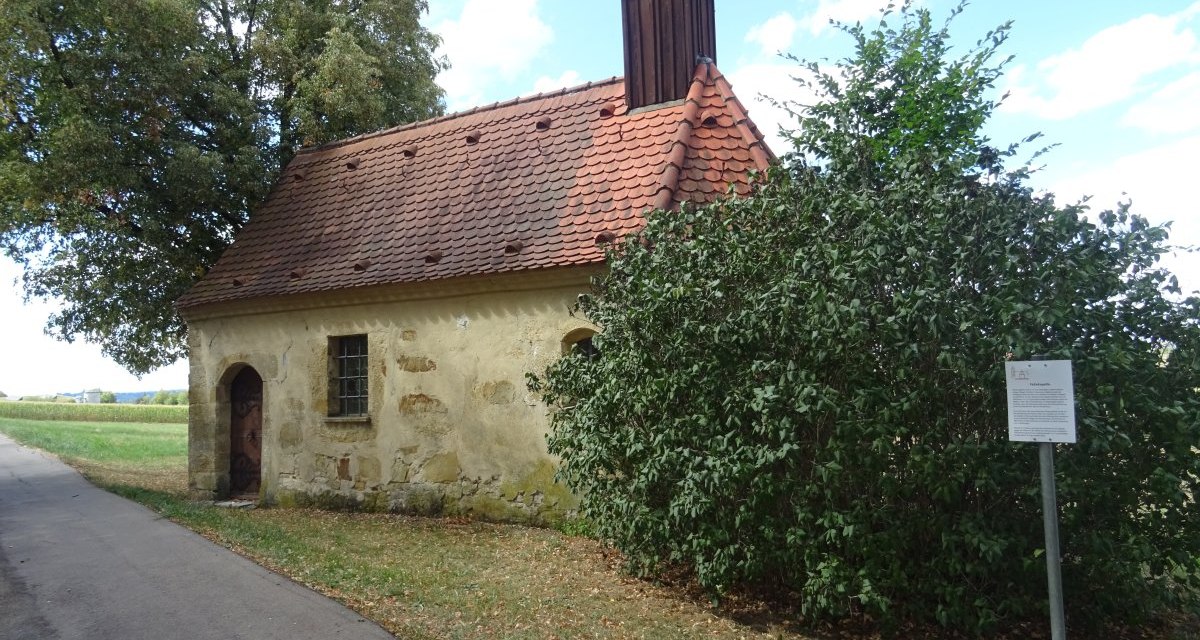 Kleine Kapelle mit rotem Ziegeldach und kleinem Turm, umgeben von Bäumen und Sträuchern. Ein Weg führt an der Kapelle vorbei., © Foto: Cornelia Steinbach Kleine Kapelle mit rotem Ziegeldach und kleinem Turm, umgeben von Bäumen und Sträuchern. Ein Weg führt an der Kapelle vorbei., © Foto: Cornelia Steinbach