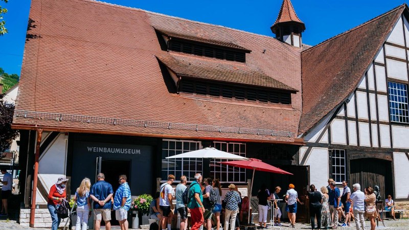 Besucher stehen vor dem Weinbaumuseum in Uhlbach. Das Fachwerkgebäude hat ein rotes Ziegeldach. Im Hintergrund sind Weinberge zu sehen., © Stuttgart-Marketing GmbH, Thomas Niedermüller Besucher stehen vor dem Weinbaumuseum in Uhlbach. Das Fachwerkgebäude hat ein rotes Ziegeldach. Im Hintergrund sind Weinberge zu sehen., © Stuttgart-Marketing GmbH, Thomas Niedermüller