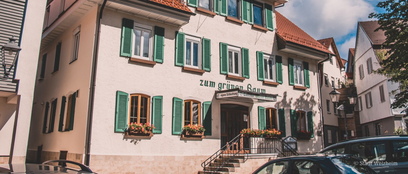 Ein traditionelles Gasthaus mit grünen Fensterläden und roten Dachziegeln, umgeben von anderen Gebäuden. Der Himmel ist blau mit einigen Wolken., © Stadt Welzheim