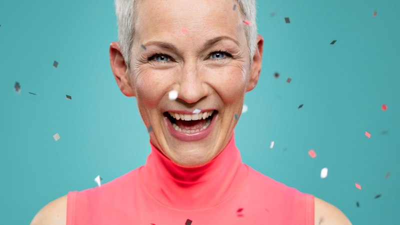 Lachende Frau mit kurzen, grauen Haaren in pinkem Oberteil vor blauem Hintergrund, umgeben von Konfetti., &copy; Tina H&auml;ussermann