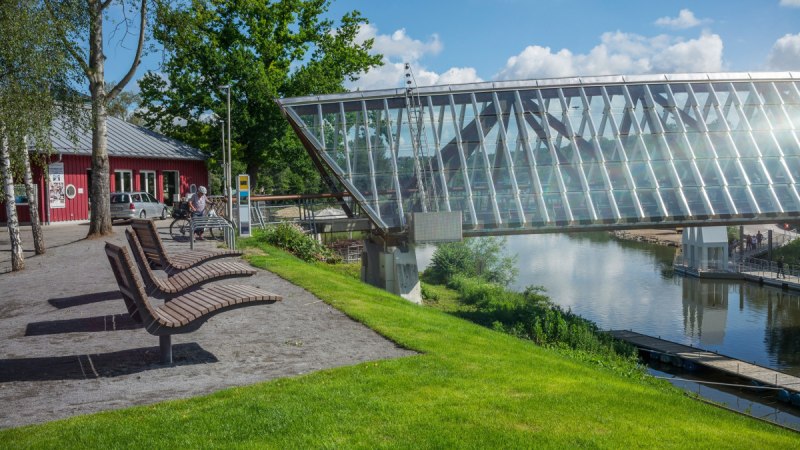 Moderne Glasbrücke über Fluss in Remseck am Neckar, umgeben von grüner Landschaft und einem roten Gebäude. Sitzbänke und Radfahrer im Vordergrund., © Remstal Tourismus e.V. Moderne Glasbrücke über Fluss in Remseck am Neckar, umgeben von grüner Landschaft und einem roten Gebäude. Sitzbänke und Radfahrer im Vordergrund., © Remstal Tourismus e.V.