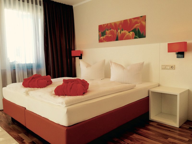 Modernes Hotelzimmer mit Doppelbett, roten Akzenten, Bild von Blumen an der Wand und zwei roten Bademänteln auf dem Bett., © TOMAS