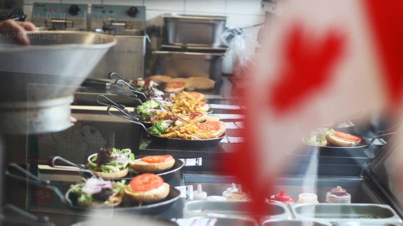 In einer Küche werden Burger und Pommes in Pfannen zubereitet. Im Vordergrund ist unscharf eine kanadische Flagge zu sehen., © SMG In einer Küche werden Burger und Pommes in Pfannen zubereitet. Im Vordergrund ist unscharf eine kanadische Flagge zu sehen., © SMG