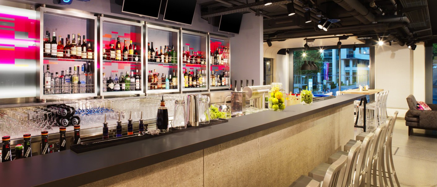 Moderne Bar mit beleuchtetem WXYZ-Schild, gut best&uuml;cktem Regal und stilvollen Sitzgelegenheiten. Die Atmosph&auml;re ist einladend und urban., &copy; Aloft Stuttgart