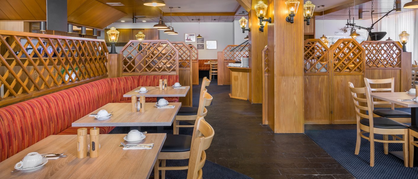 Gemütlicher Frühstücksbereich mit Holzmöbeln, roten Polstern und gedeckten Tischen. Warme Beleuchtung sorgt für eine einladende Atmosphäre., © Novum Management GmbH