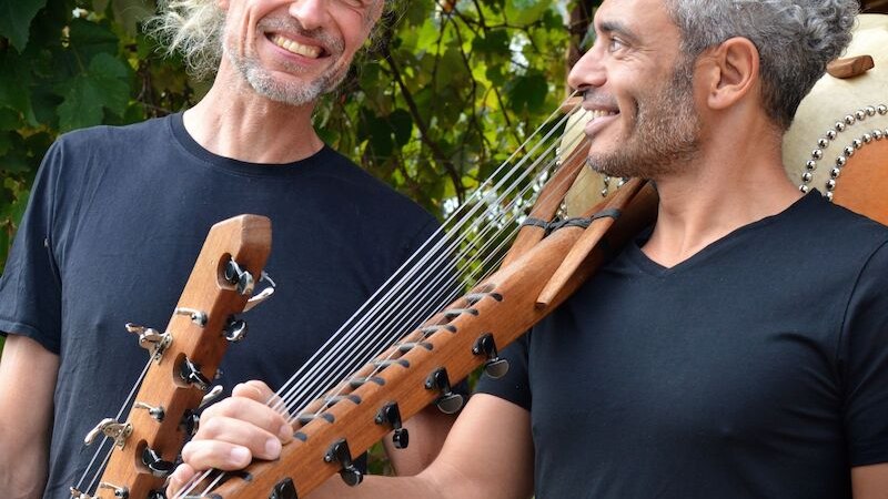 Zwei Männer mit Saiteninstrumenten lächeln sich an. Sie stehen vor einer grünen, blättrigen Kulisse und tragen schwarze T-Shirts., © Interessengemeinschaft Kultur Sindelfingen / Böblingen e.V. Zwei Männer mit Saiteninstrumenten lächeln sich an. Sie stehen vor einer grünen, blättrigen Kulisse und tragen schwarze T-Shirts., © Interessengemeinschaft Kultur Sindelfingen / Böblingen e.V.