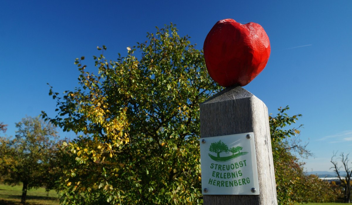 Ein Holzpfosten mit rotem Apfel und Schild 'Streuobst Erlebnis Herrenberg' vor blauem Himmel und B&auml;umen., &copy; Natur.Nah. Sch&ouml;nbuch & Heckeng&auml;u
