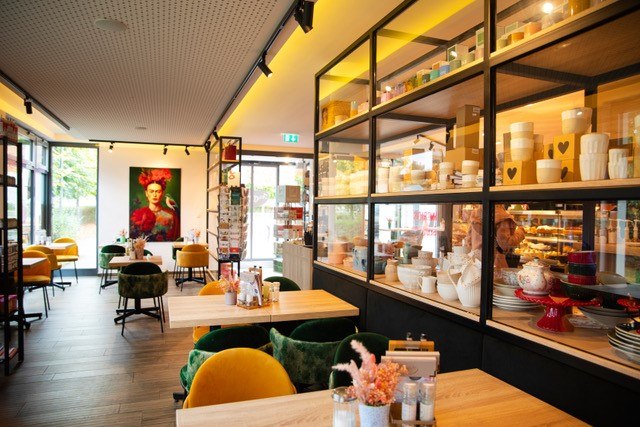 Gemütliches Café mit Holztischen, bunten Stühlen und Geschirrregalen. Ein buntes Porträt schmückt die Wand., © Ingrid Glückswelt