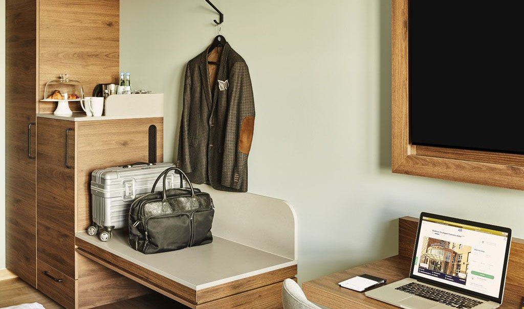 Ein modernes Hotelzimmer mit einem Laptop auf dem Schreibtisch, einem Koffer und einer Jacke an der Wand. Ein Schrank mit Getränken und Snacks., © Pullman Stuttgart Fontana Ein modernes Hotelzimmer mit einem Laptop auf dem Schreibtisch, einem Koffer und einer Jacke an der Wand. Ein Schrank mit Getränken und Snacks., © Pullman Stuttgart Fontana