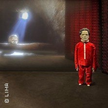 Ein puppenartiges Wesen in rotem Anzug steht in einem dunklen Raum. Im Hintergrund erscheint ein Gesicht im Lichtstrahl., &copy; links im Bild