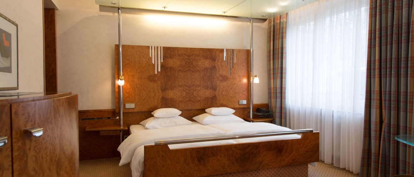 Elegantes Hotelzimmer mit Doppelbett, Holzmöbeln und großen Fenstern mit Vorhängen. Ein Teller mit Obst steht auf einem Glastisch., © Hotel Royal Elegantes Hotelzimmer mit Doppelbett, Holzmöbeln und großen Fenstern mit Vorhängen. Ein Teller mit Obst steht auf einem Glastisch., © Hotel Royal