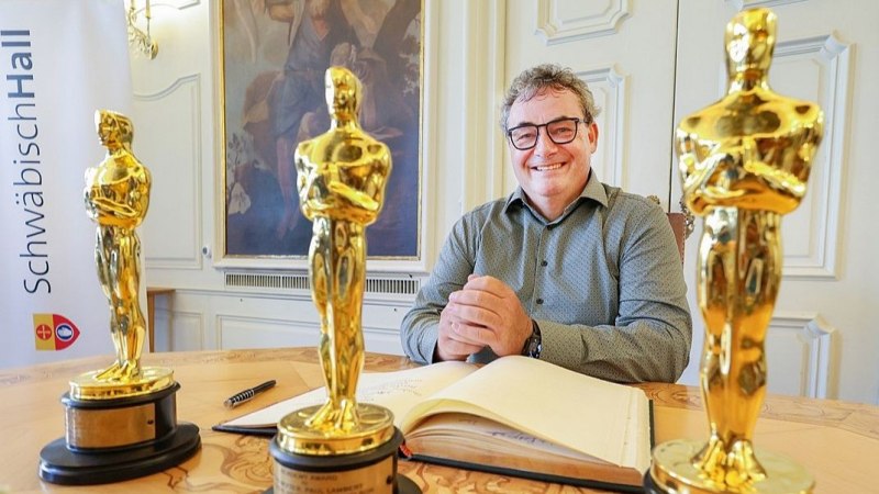 Ein Mann sitzt l&auml;chelnd an einem Tisch mit drei Oscar-Statuetten. Im Hintergrund ist ein Gem&auml;lde und ein Banner mit der Aufschrift 'Schw&auml;bisch Hall' zu sehen., &copy; S&uuml;dwest Presse Hohenlohe