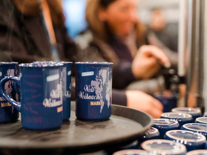 Blaue Tassen mit 'Welt Weihnachts Markt' stehen auf einem Tablett. Im Hintergrund schenken Menschen Glühwein ein, was eine festliche Atmosphäre schafft., © Cool-Tours StattReisen