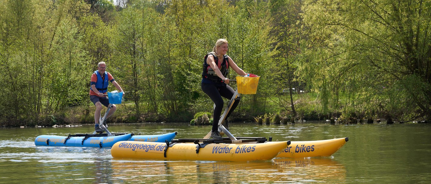Zwei Personen fahren auf gelben Wasserfahrrädern auf einem Fluss. Sie tragen Schwimmwesten und sind von grüner Natur umgeben., © Foto LKZ - Fotograf Andreas Becker