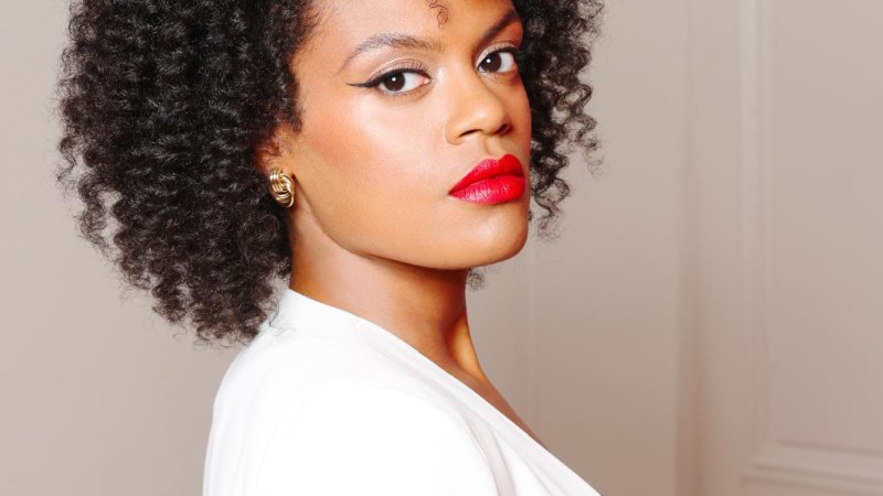 Frau mit lockigem Haar und rotem Lippenstift in weißem Mantel, seitlich blickend., © Ludovic Baron Frau mit lockigem Haar und rotem Lippenstift in weißem Mantel, seitlich blickend., © Ludovic Baron