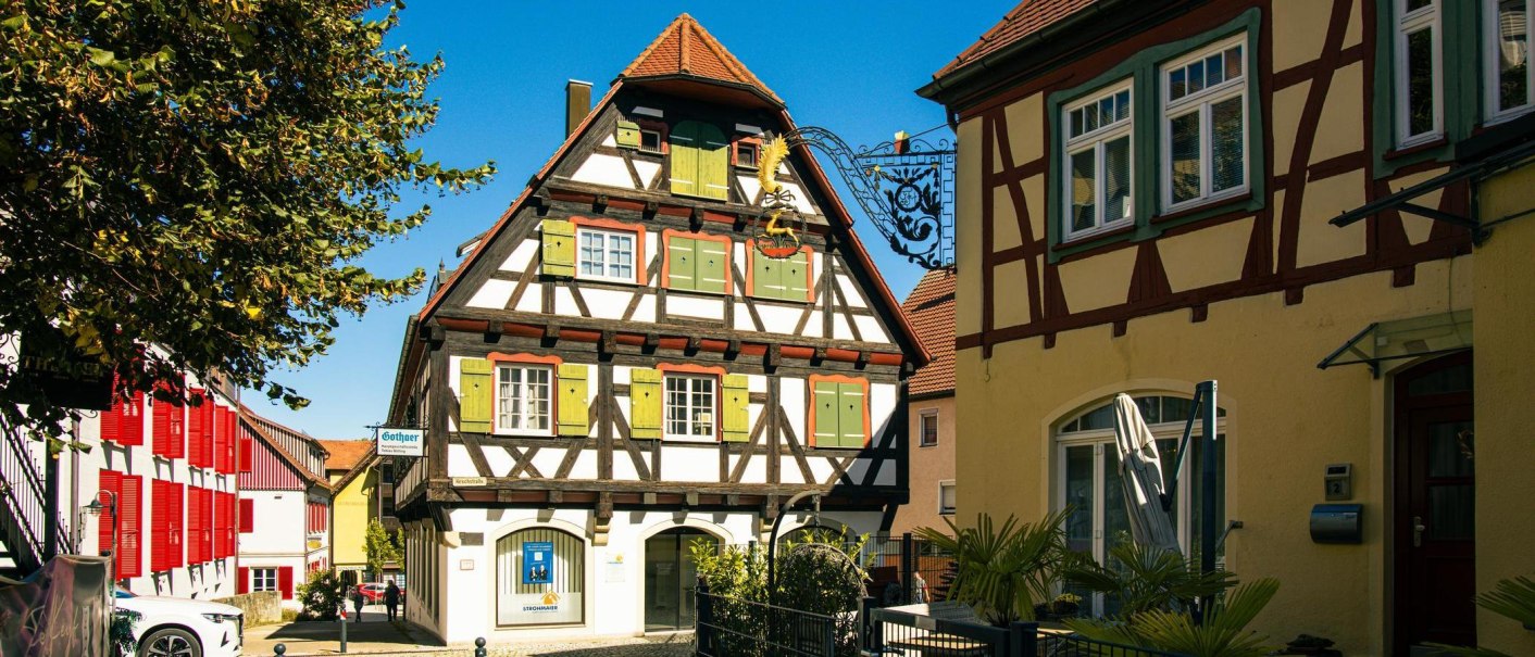 Fachwerkhaus in Gaildorf mit grünen Fensterläden und rotem Dach, umgeben von weiteren historischen Gebäuden und einem Baum im Vordergrund., © Stuttgart-Marketing GmbH, Sarah Schmid