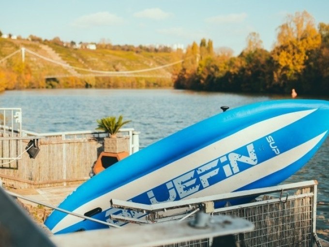 Blau-weißes SUP-Board am Flussufer, Hängebrücke und herbstliche Landschaft im Hintergrund., © Cool-Tours StattReisen Blau-weißes SUP-Board am Flussufer, Hängebrücke und herbstliche Landschaft im Hintergrund., © Cool-Tours StattReisen
