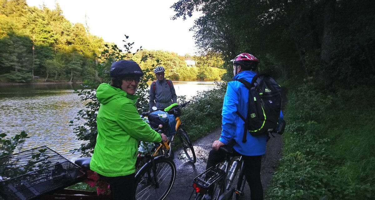 Drei Radfahrer pausieren am Bärensee. Sie tragen Helme und bunte Jacken, während sie die Natur genießen. Der See und Bäume bilden die Kulisse., © Natur.Nah. Schönbuch & Heckengäu Drei Radfahrer pausieren am Bärensee. Sie tragen Helme und bunte Jacken, während sie die Natur genießen. Der See und Bäume bilden die Kulisse., © Natur.Nah. Schönbuch & Heckengäu