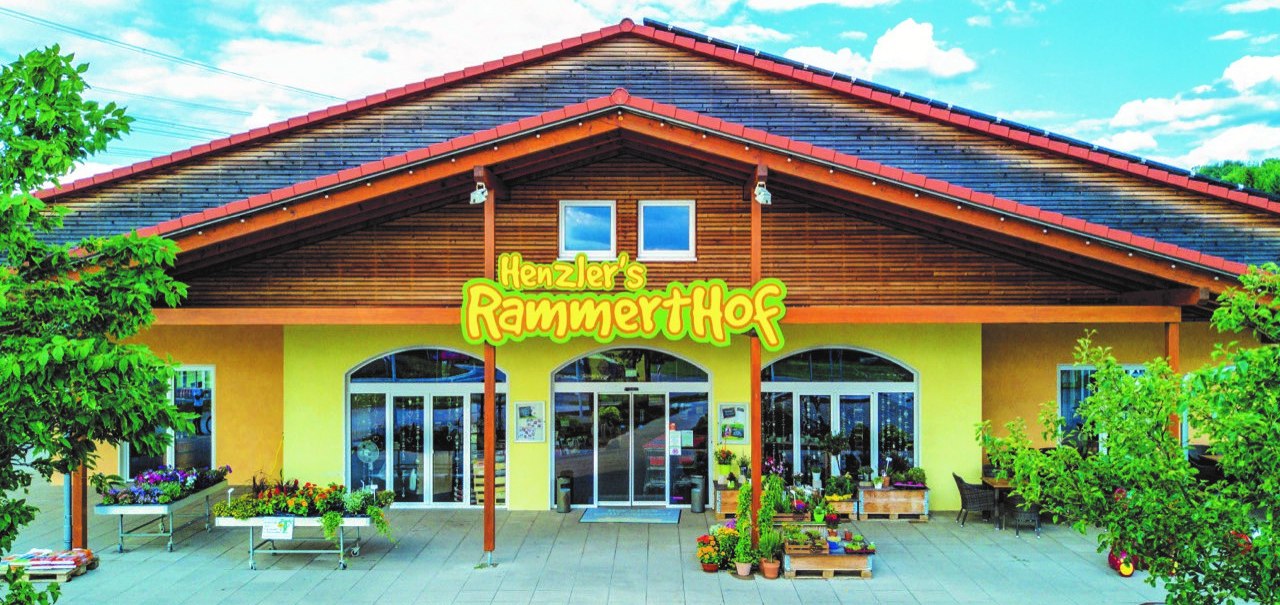 Frontansicht des Rammerthofs mit großem gelbem Schild, Pflanzen und Blumen vor dem Eingang. Das Gebäude hat ein rotes Dach und Holzelemente., © Engel/ Rammerthof