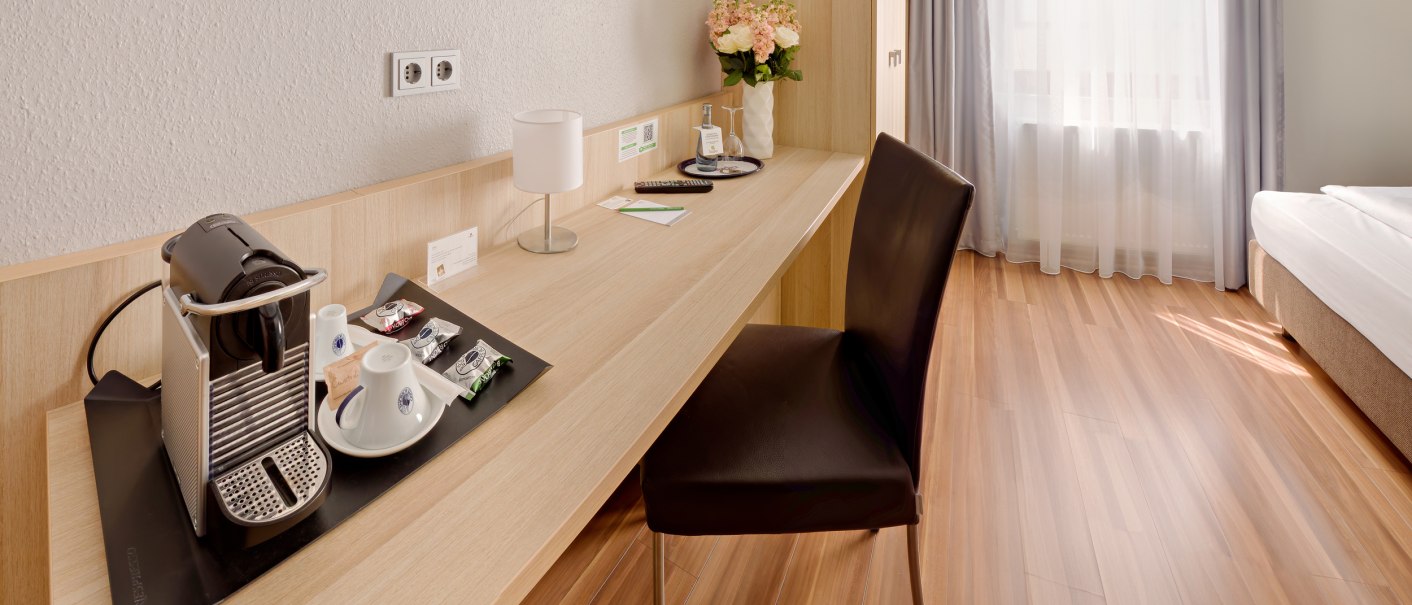 Modernes Einzelzimmer mit Holzfußboden, Schreibtisch, Stuhl, Kaffeemaschine und Bett. Helle Vorhänge lassen Tageslicht herein., © attimo Hotel Stuttgart