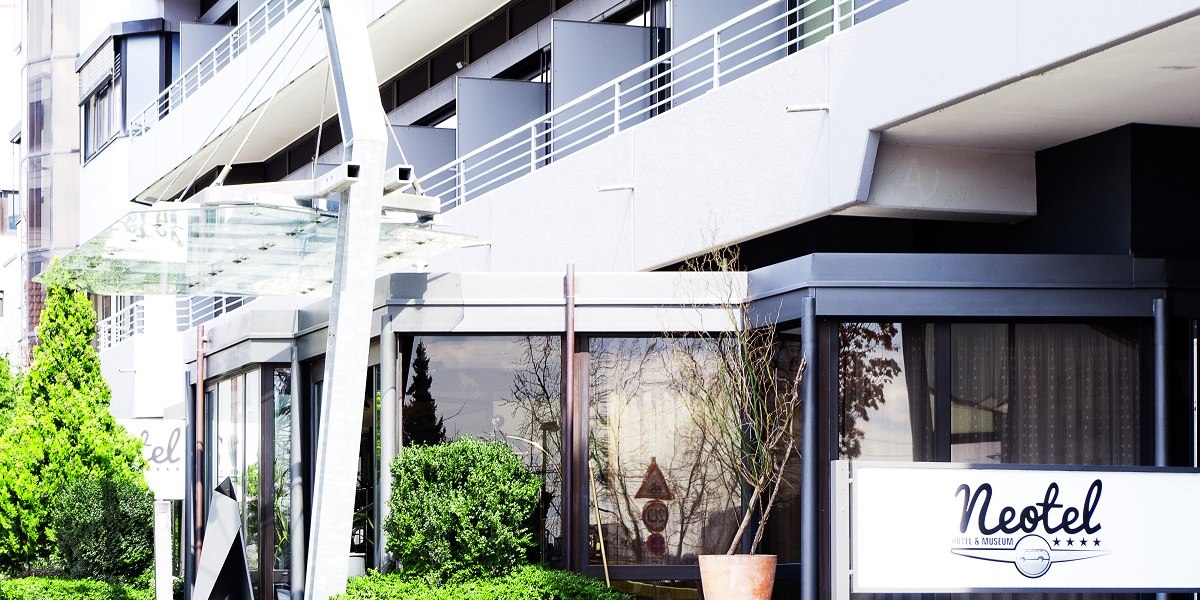 Eingang des Hotels Neotel mit Glasvordach und moderner Fassade. Ein Schild mit dem Hotelnamen ist sichtbar. Grüne Pflanzen umrahmen den Eingang., © Hotel Neotel