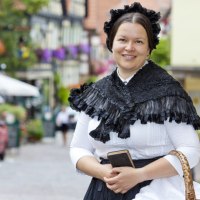 Eine Frau in historischer Kleidung des 19. Jahrhunderts steht l&auml;chelnd in einer malerischen Altstadt mit Fachwerkh&auml;usern und Blumen., &copy; Stadt Besigheim
