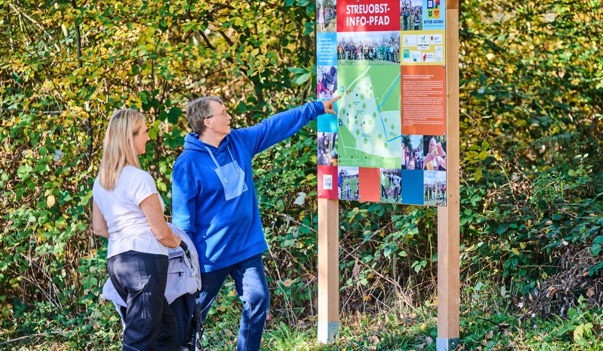 Zwei Personen stehen vor einer Informationstafel mit der Aufschrift 'Streuobst-Info-Pfad' in einem bewaldeten Gebiet., &copy; Natur.Nah. Sch&ouml;nbuch & Heckeng&auml;u