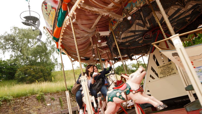 Zwei Personen fahren auf einem nostalgischen Karussell mit Pferdefiguren. Im Hintergrund ist ein spiralf&ouml;rmiger Aussichtsturm zu sehen., &copy; SMG, Christoph D&uuml;pper