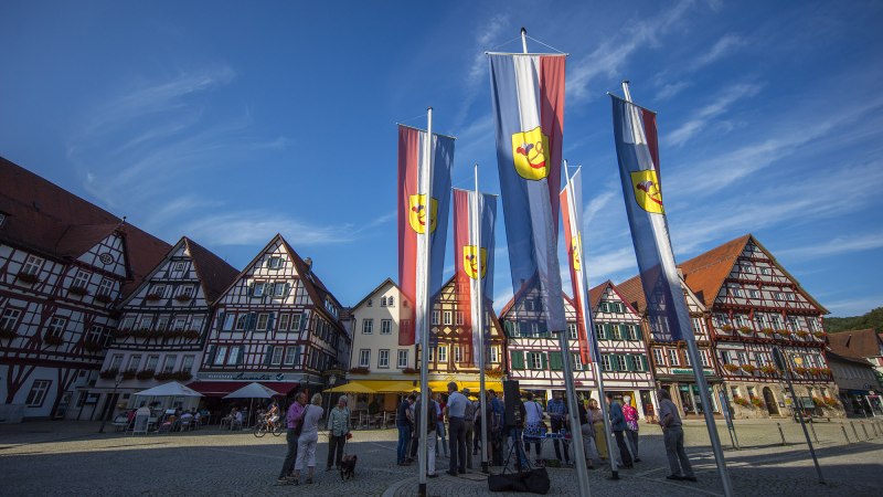 Der Marktplatz von Bad Urach zeigt Fachwerkh&auml;user und Fahnen. Menschen stehen auf dem gepflasterten Platz unter blauem Himmel., &copy; Bad Urach Tourismus