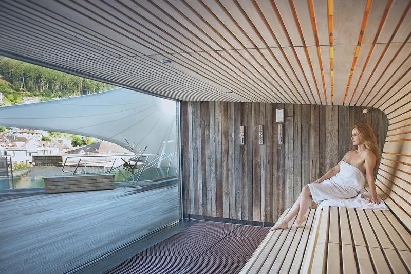 Eine Frau sitzt in einer modernen Sauna mit Holzverkleidung im Palais Thermal. Durch die Glaswand sieht man eine Terrasse und grüne Hügel., © Mokni's Palais Hotel & Spa Eine Frau sitzt in einer modernen Sauna mit Holzverkleidung im Palais Thermal. Durch die Glaswand sieht man eine Terrasse und grüne Hügel., © Mokni's Palais Hotel & Spa