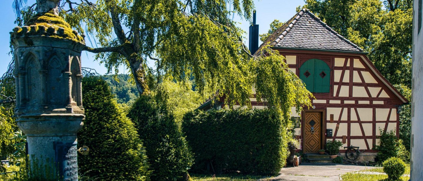 Ein malerisches Fachwerkhaus mit grünem Tor, umgeben von üppigem Grün und einem alten Steinmonument im Vordergrund., © Stuttgart-Marketing GmbH, Sarah Schmid Ein malerisches Fachwerkhaus mit grünem Tor, umgeben von üppigem Grün und einem alten Steinmonument im Vordergrund., © Stuttgart-Marketing GmbH, Sarah Schmid